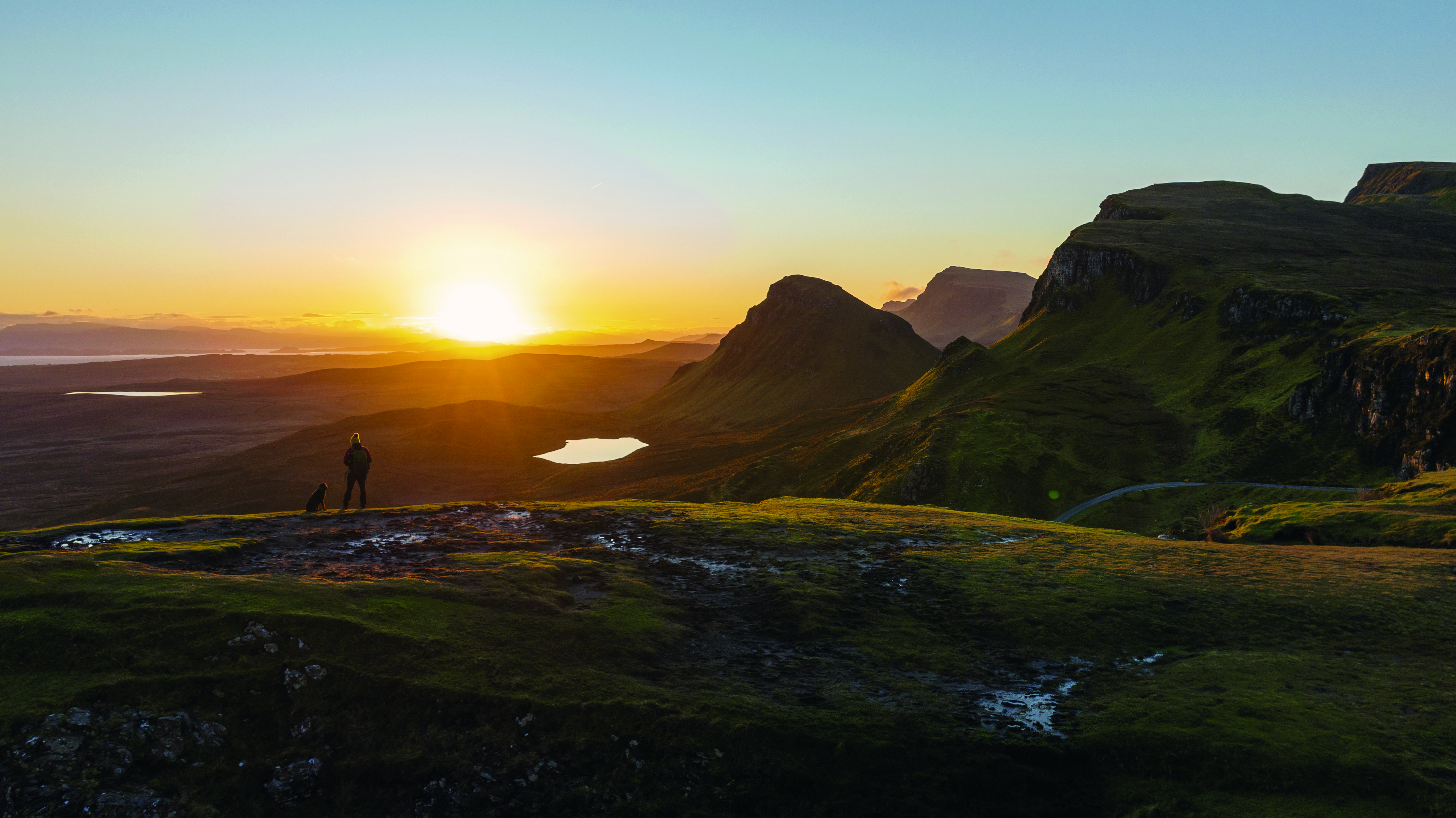 An image taken at sunset of a beautiful, green landscape. A person and a dog are in the distance looking out into the distance.